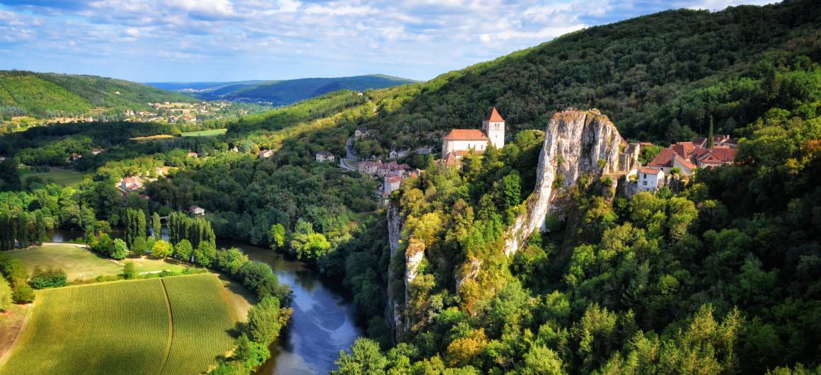 Scenic view of the Lot River at the base of tall limestone cliffs, surrounded by lush greenery.