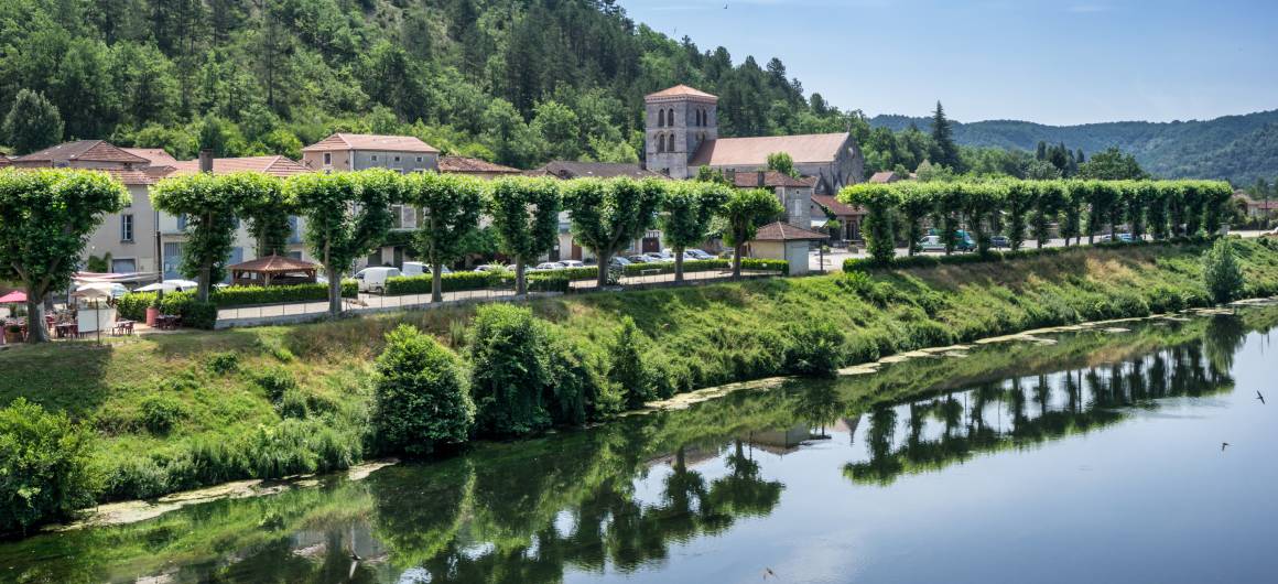 Charming riverside village in the Lot with neatly trimmed trees along the bank and wooded hills in the background.