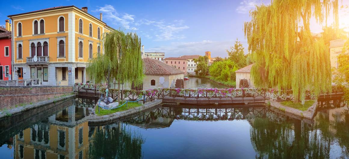 Charming canal in Treviso with weeping willows and a flower‑covered bridge glowing in the warm evening light.