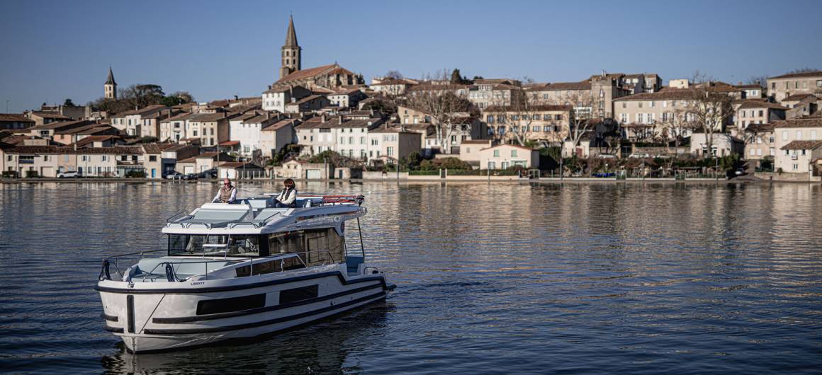 A Liberty boat cruising peacefully on a basin facing the city of Castelnaudary, with its houses and church tower in the background under a clear sky.