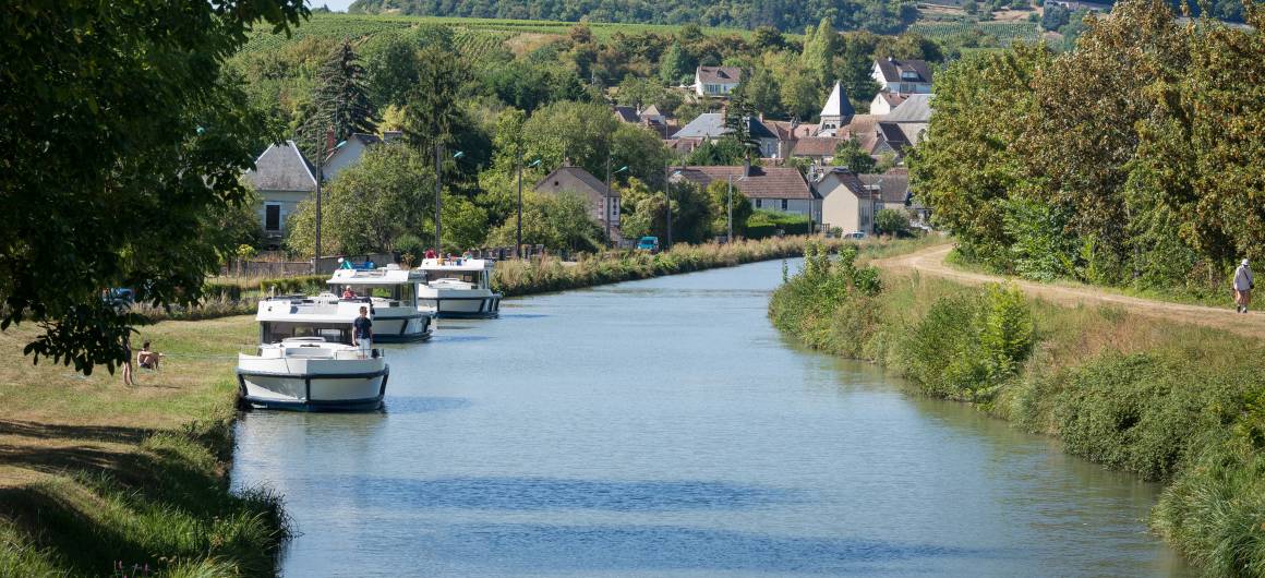 Canal boats cruising past a charming village in Burgundy, with vineyards and houses nestled in the green hills.