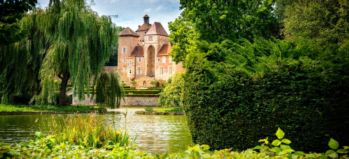 Un gran castillo de arquitectura clásica francesa está parcialmente oculto tras los árboles, junto a un apacible río.