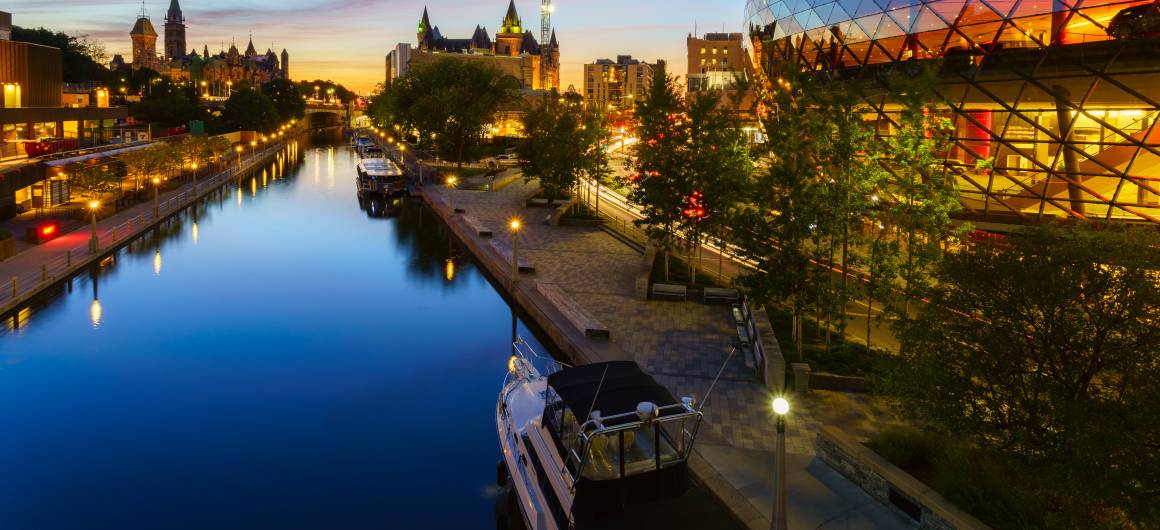 Evening lights glow along the Ottawa city waterfront as boats line the dock on the Rideau Canal beneath the downtown skyline.