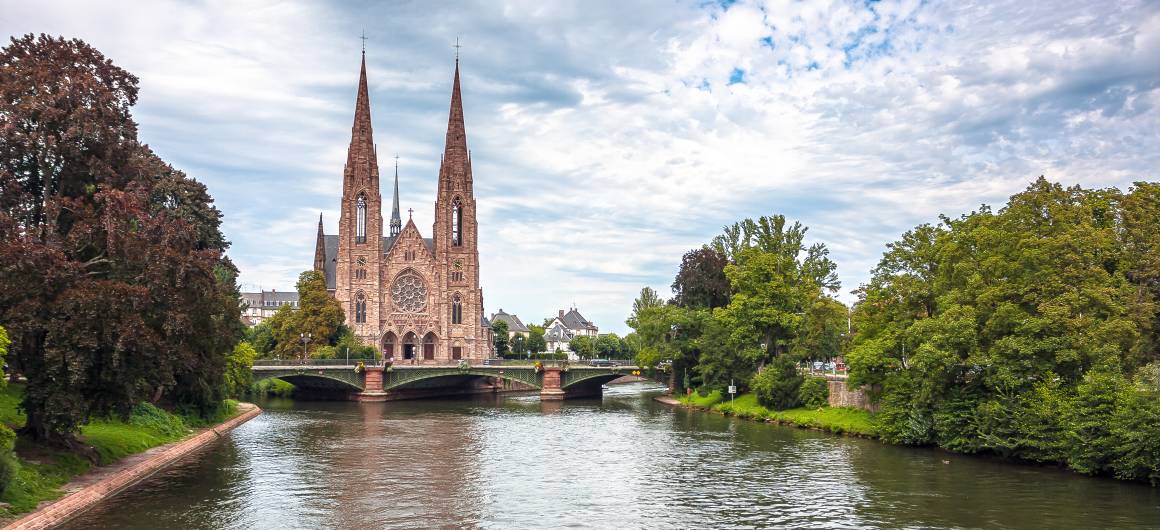 Église Saint‑Paul à Strasbourg se reflétant dans le fleuve, encadrée par de riches feuillages et un ciel d’été nuageux.