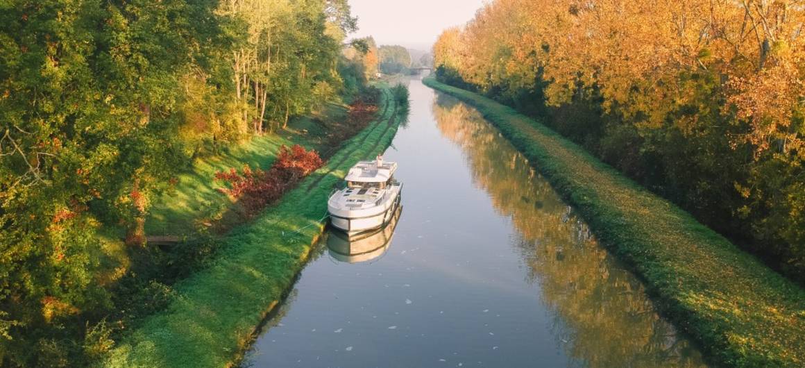 A boat cruises down a tree-lined canal in Burgundy during autumn, with golden foliage reflecting in the calm water.