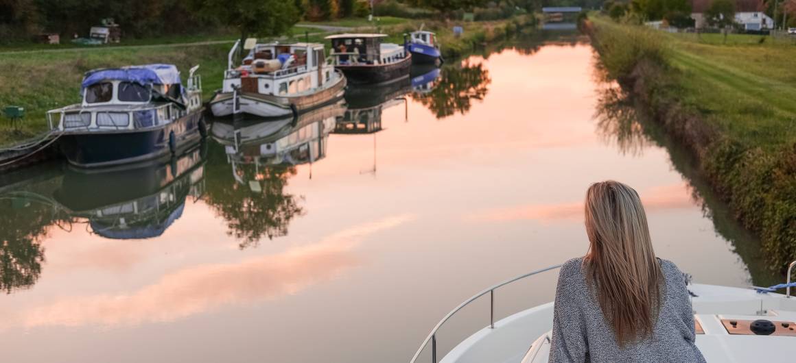 A woman sits at the front of a boat at sunset, gazing over calm canal waters with moored boats nearby in Burgundy.