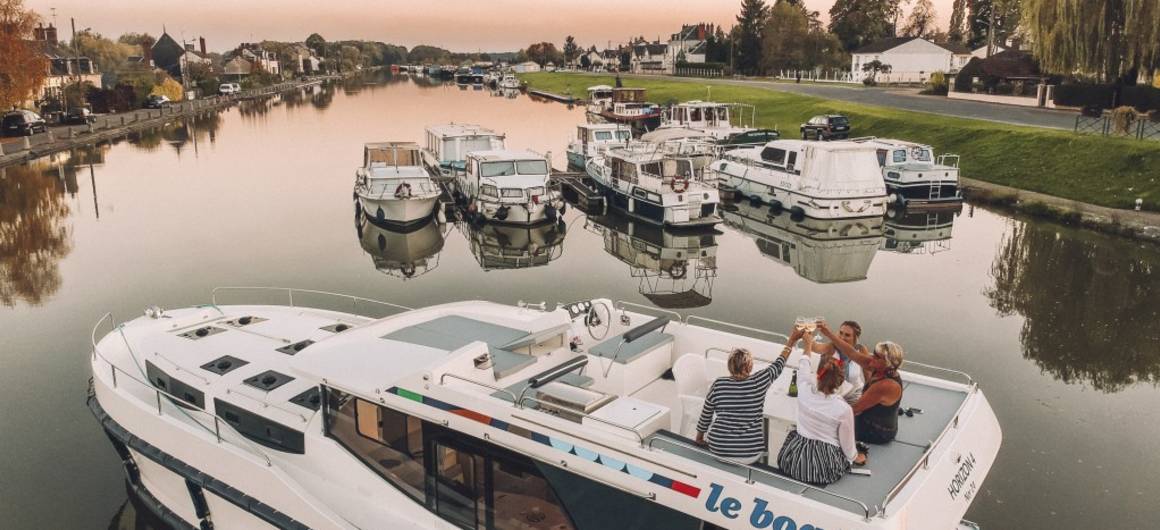 A group of friends raise glasses aboard a moored boat at sunset, with a marina and other boats in the background.
