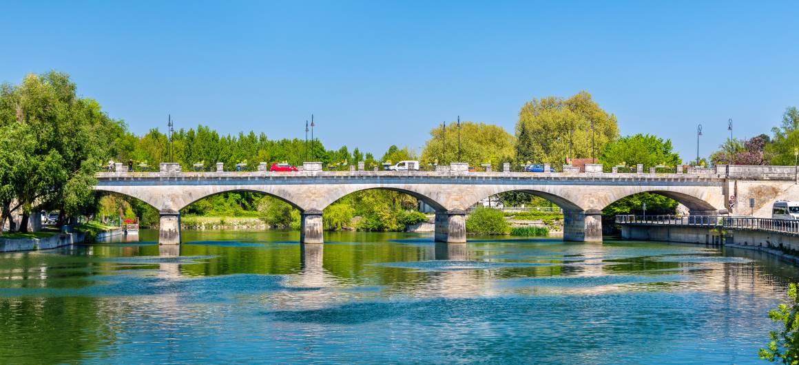 Uitzicht op de Pont Neuf in Cognac die de Charente oversteekt, met auto's en voetgangers die over het kalme water passeren.
