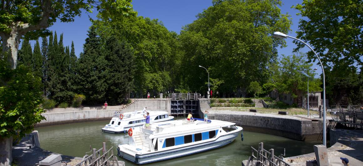 A canal boat navigates toward a traditional lock on the Canal du Midi in Agde, a hallmark of canal cruising in France.
