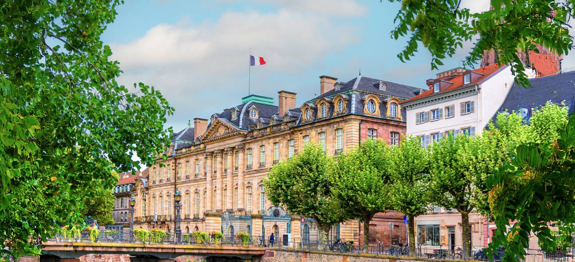 Vue sur le grand Palais Rohan à Saverne, avec le drapeau français flottant au-dessus et des arbres feuillus longeant le canal.