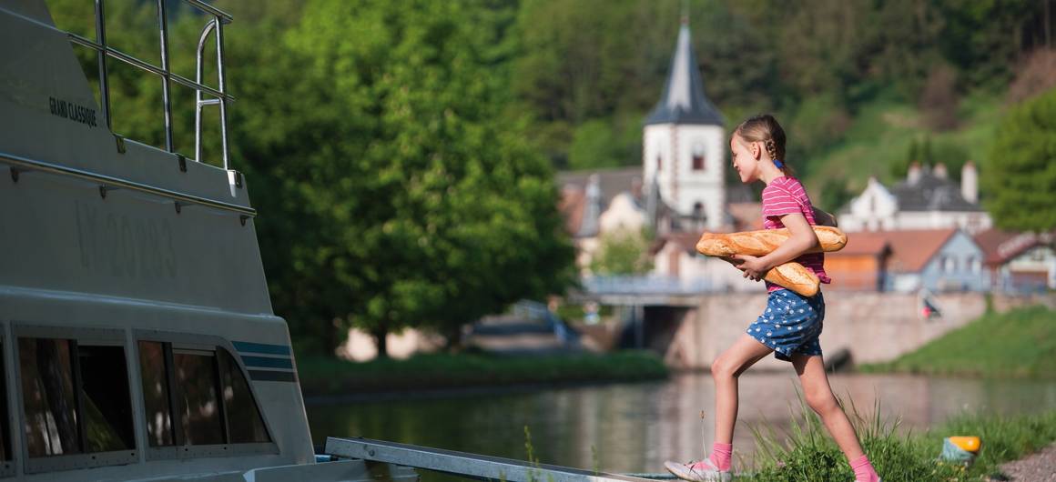 Jeune fille portant des baguettes en montant à bord d’un bateau amarré près d’un village en Alsace.