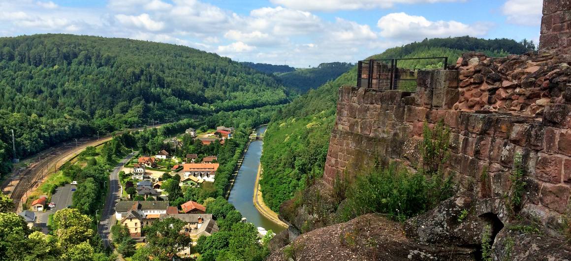 Vue aérienne depuis les ruines du château de Lutzelbourg, surplombant le canal de la Marne‑Rhine serpentant à travers une vallée boisée et les toits d’un village.