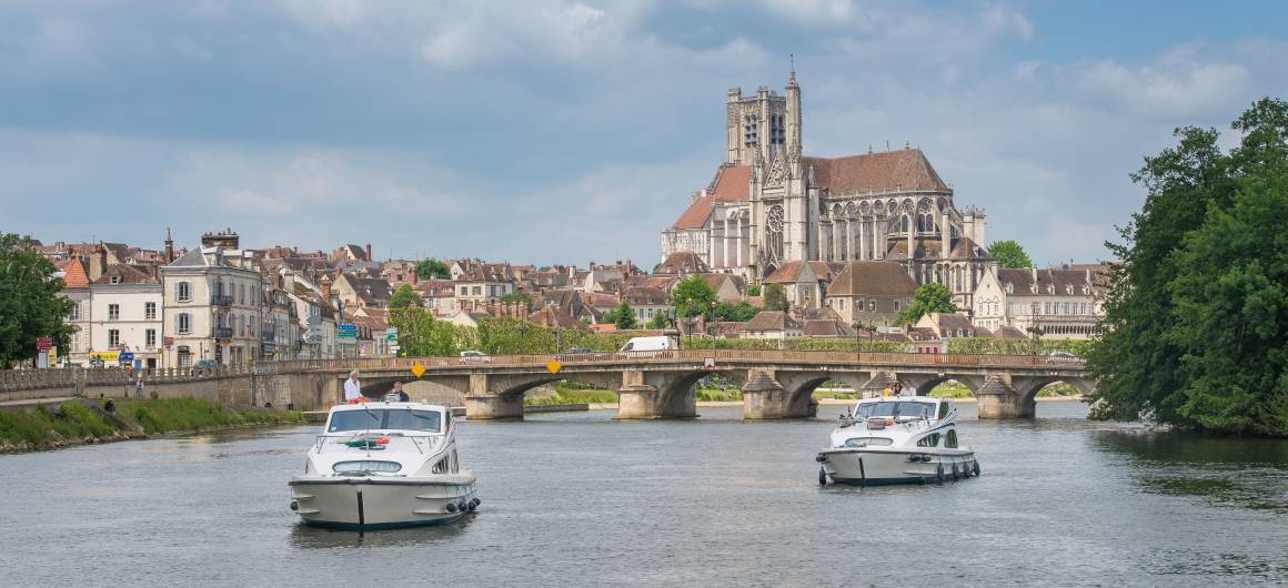 Boats approach a historic Burgundian town with a majestic cathedral and stone bridge spanning the river.