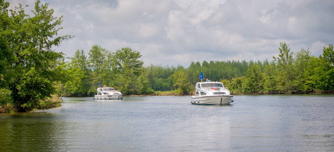 Two canal boats cruise peacefully through open waters under blue skies, surrounded by greenery.