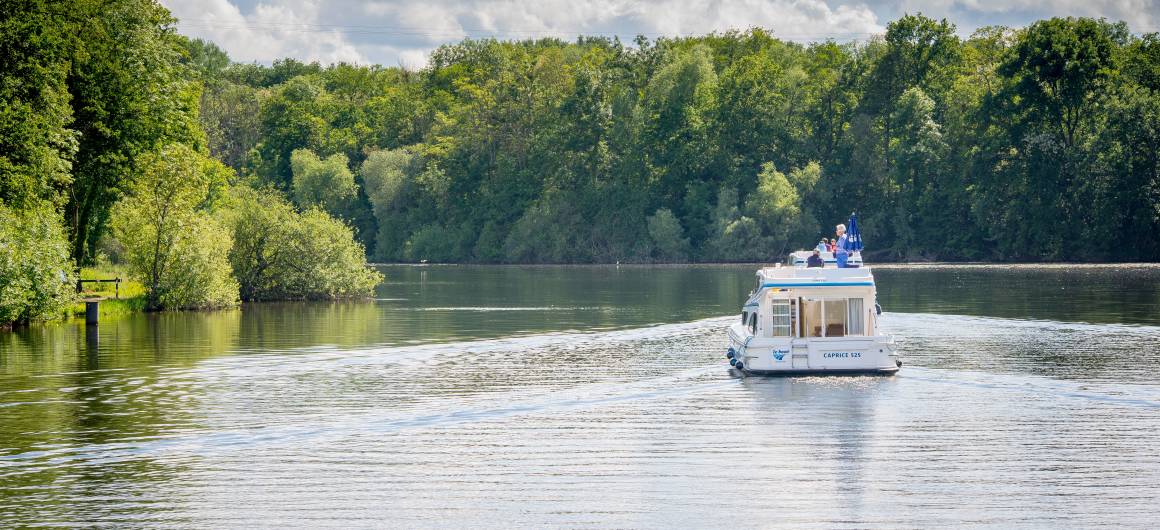 A canal boat cruises through a wide river bordered by dense green forest under a partly cloudy sky.