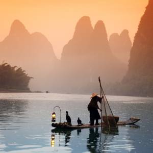 Silhouette eines Fischers und Kormoranen auf dem Li-Fluss in Yangshuo, China, im Hintergrund das Karstgebirge im Licht des Sonnenuntergangs
