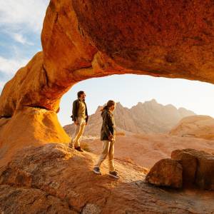 Zwei Personen unter einem Felsbogen bei Sonnenaufgang im Spitzkoppe-Gebiet im Damaraland, Namibia