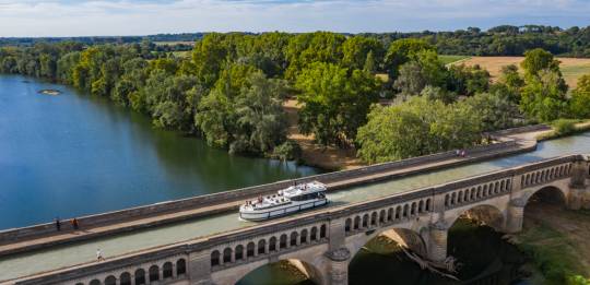 A canal boat cruises across the Orb Aqueduct on the Canal du Midi, surrounded by peaceful countryside.