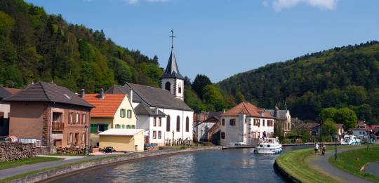 Boote befahren den Kanal im hessischen Dorf mit bunten Häusern, einer Kirche und grünen Hügeln unter hellem Himmel.