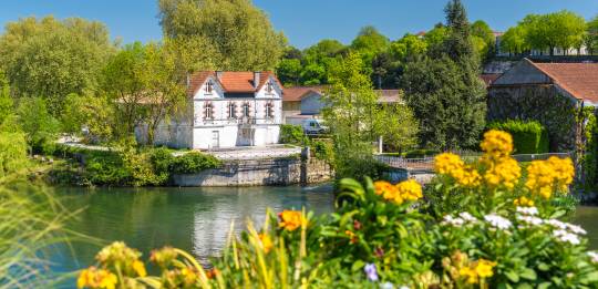 Charming riverside house in Charente surrounded by blooming flowers and lush greenery under a vibrant blue sky.