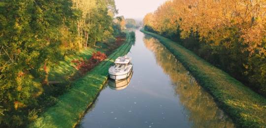 A boat cruises down a tree-lined canal in Burgundy during autumn, with golden foliage reflecting in the calm water.