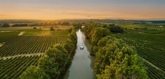 A lone canal boat sails into the golden sunset on the Canal du Midi, cutting through vineyards and rural countryside for a picture-perfect moment on a French boating holiday.