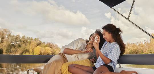 Une mère et sa fille rient en jouant aux cartes sur le bateau, entourées d'arbres dorés d'automne et d'un ciel calme.