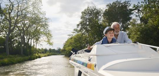 Grandfather and grandson steering a canal boat together on a sunny countryside journey.