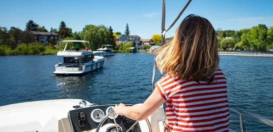 Young woman at helm of Le Boat Horizon houseboat during approach to Westport Marina on Big Rideau Lake, Westport, Ontario, Canada