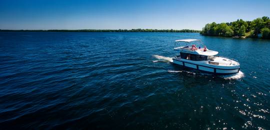 Aerial view of a Le Boat cruiser gliding along the calm Rideau Canal in Canada, surrounded by lush green forests under a clear blue sky.