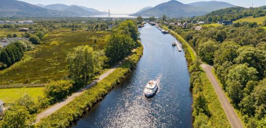 Aerial view of a motorboat cruising along a calm Scottish canal, surrounded by green countryside and hills