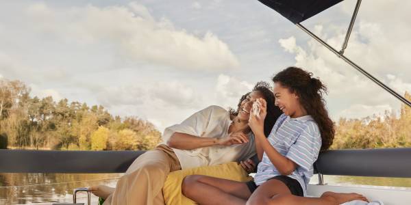 Eine Mutter und ihre Tochter brechen in Lachen aus, während sie zusammen auf dem Boot Karten spielen, umgeben von goldenen Herbstbäumen und einem wunderschönen Himmel.