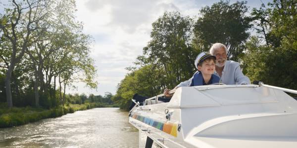 Grandfather and grandson steering a houseboat together on a sunny countryside journey.