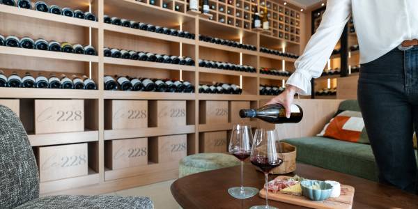 Wine lounge with shelves of bottles, a table with two glasses of red wine, and a plate of cheese and charcuterie.