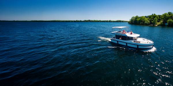 Aerial view of a Le Boat cruiser gliding along the calm Rideau Canal in Canada, surrounded by lush green forests under a clear blue sky.