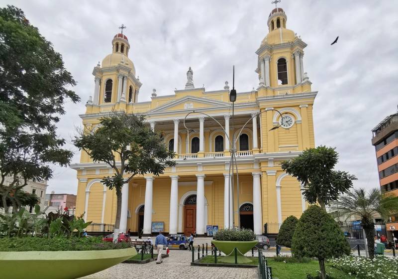 Yellow colonial cathedral with twin domed bell towers and white columns, fronted by a park with trimmed trees in Chiclayo, Peru.