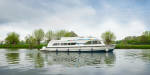 Side view of the Dancer 3 gliding along a peaceful river, mirrored on the water’s surface, with soft clouds and spring foliage lining the banks.