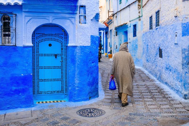 Person in a traditional djellaba walking through a narrow blue-painted alley in Chefchaouen's medina, Morocco.