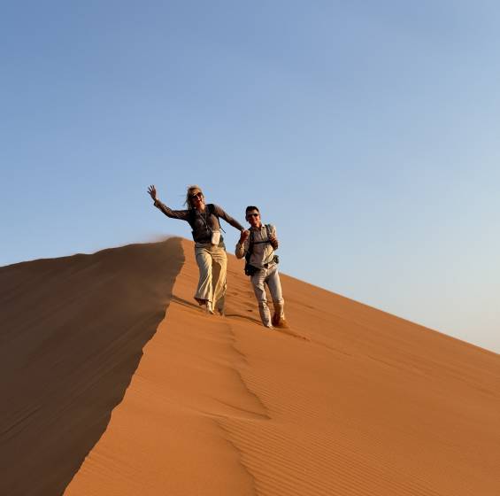 Zwei fröhliche Reisende laufen eine rote Sanddüne in der namibischen Wüste hinunter, blauer Himmel im Hintergrund.