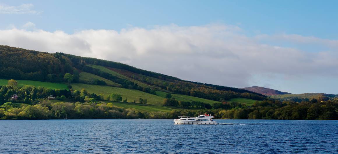 A Le Boat's cruiser sailing across a loch, backed by patchworked green and forested hills under a sunny sky.