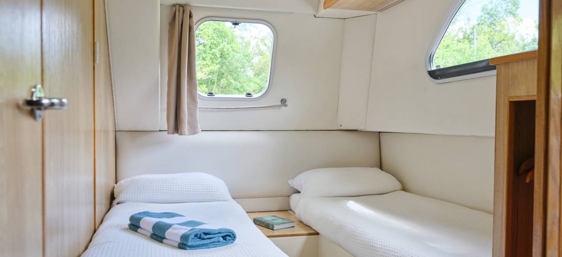Twin cabin inside the Dancer 4 with crisp white bedding, wooden walls, and views of greenery through the windows.