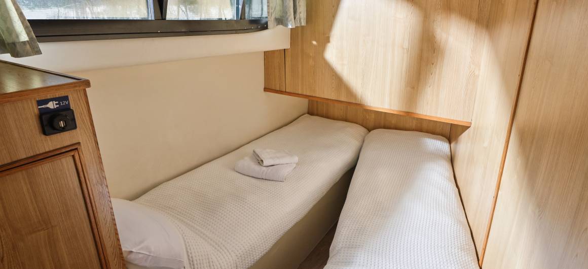Compact twin cabin aboard the Elegance, with two simple beds along wood-panelled walls, bathed in warm natural light through curtained windows.
