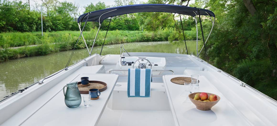 Top deck of the Vision 4 with dining area, striped towel, apples, and a view of calm canal waters.