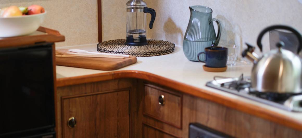 Wood-finished kitchen area on the Grand Classique with a gas hob, kettle, French press, water jug, and fruit bowl set along a light worktop.