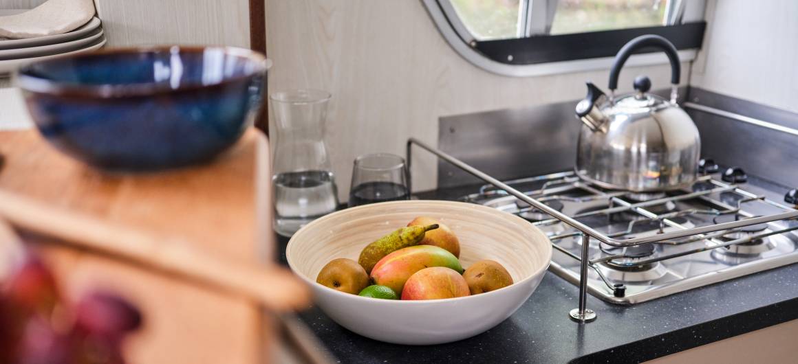 Modern kitchen setup in the Continentale with a shiny kettle on the stove, black countertop, and a fresh fruit bowl beside a window.