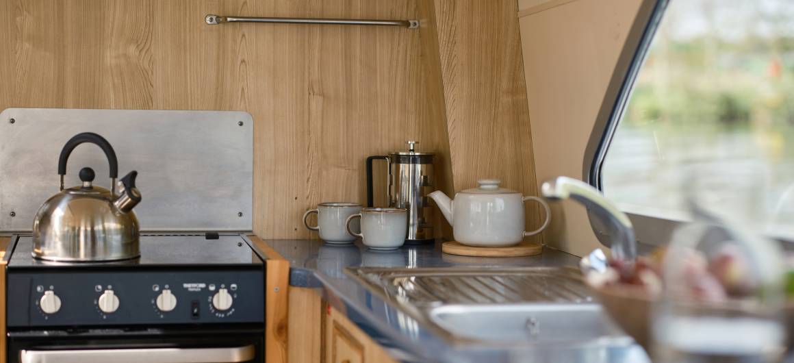 Compact galley kitchen aboard the Dancer 2, featuring a kettle on the stove, mugs, and a teapot neatly set up by the window overlooking the river.