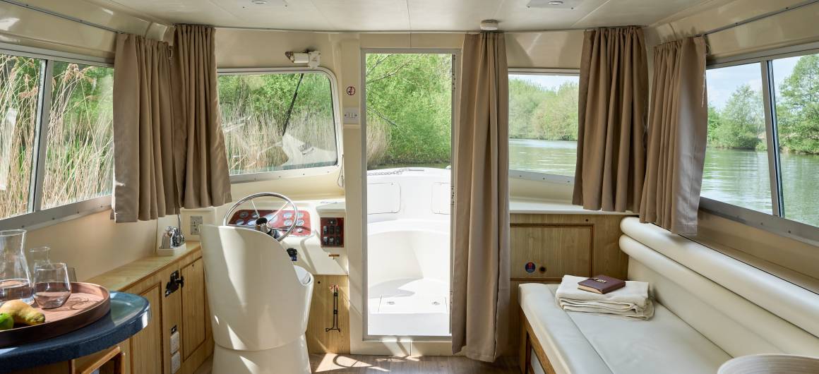 Bright saloon on the Dancer 2 with white leather seating, a steering console, and a door to the deck, framed by large windows with lush riverside views.