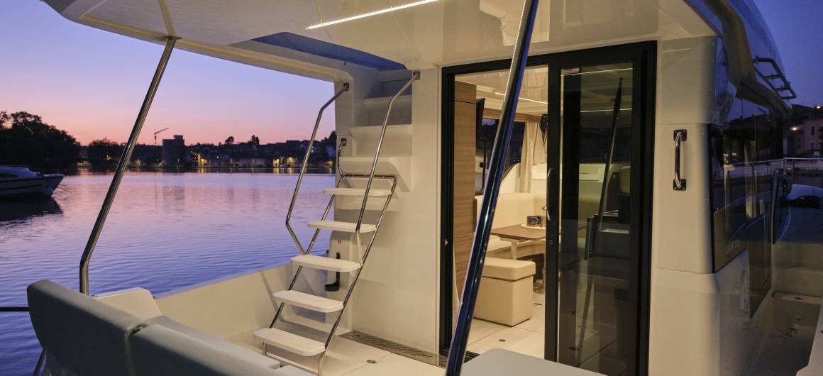 Exterior stern view of the Liberty boat at sunset, showing steps leading to the top deck, and a glimpse into the illuminated saloon through glass doors.