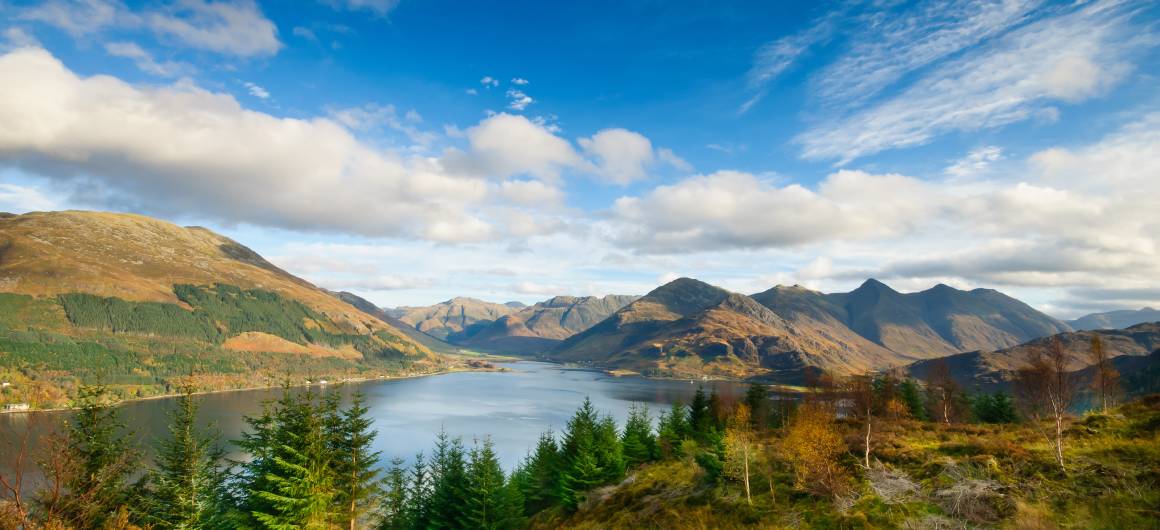 Ein Panoramablick auf einen schottischen See, flankiert von sanften Hügeln und zerklüfteten Bergen unter einem hellblauen Himmel mit verstreuten Wolken.