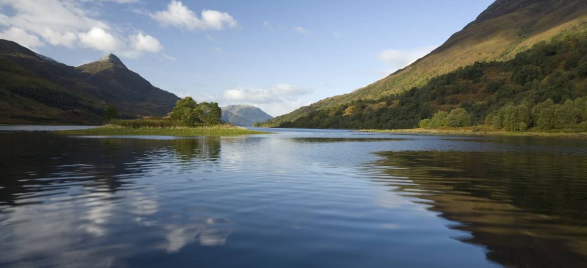 Ein weiter Loch, flankiert von Highlands-Bergen und bewaldeten Hängen, mit weichen Wolkenspiegelungen im stillen Wasser.